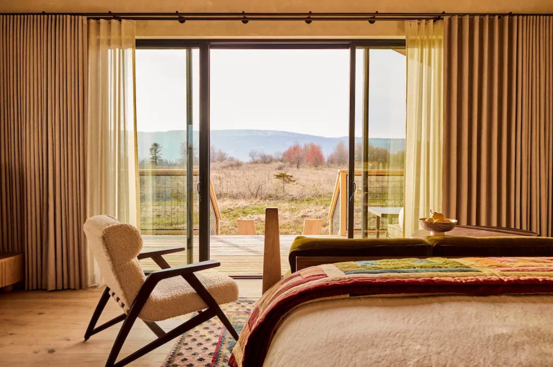 Cozy bedroom with sliding glass doors revealing a serene landscape of fields and distant mountains. Warm colors and natural light create a peaceful ambiance.