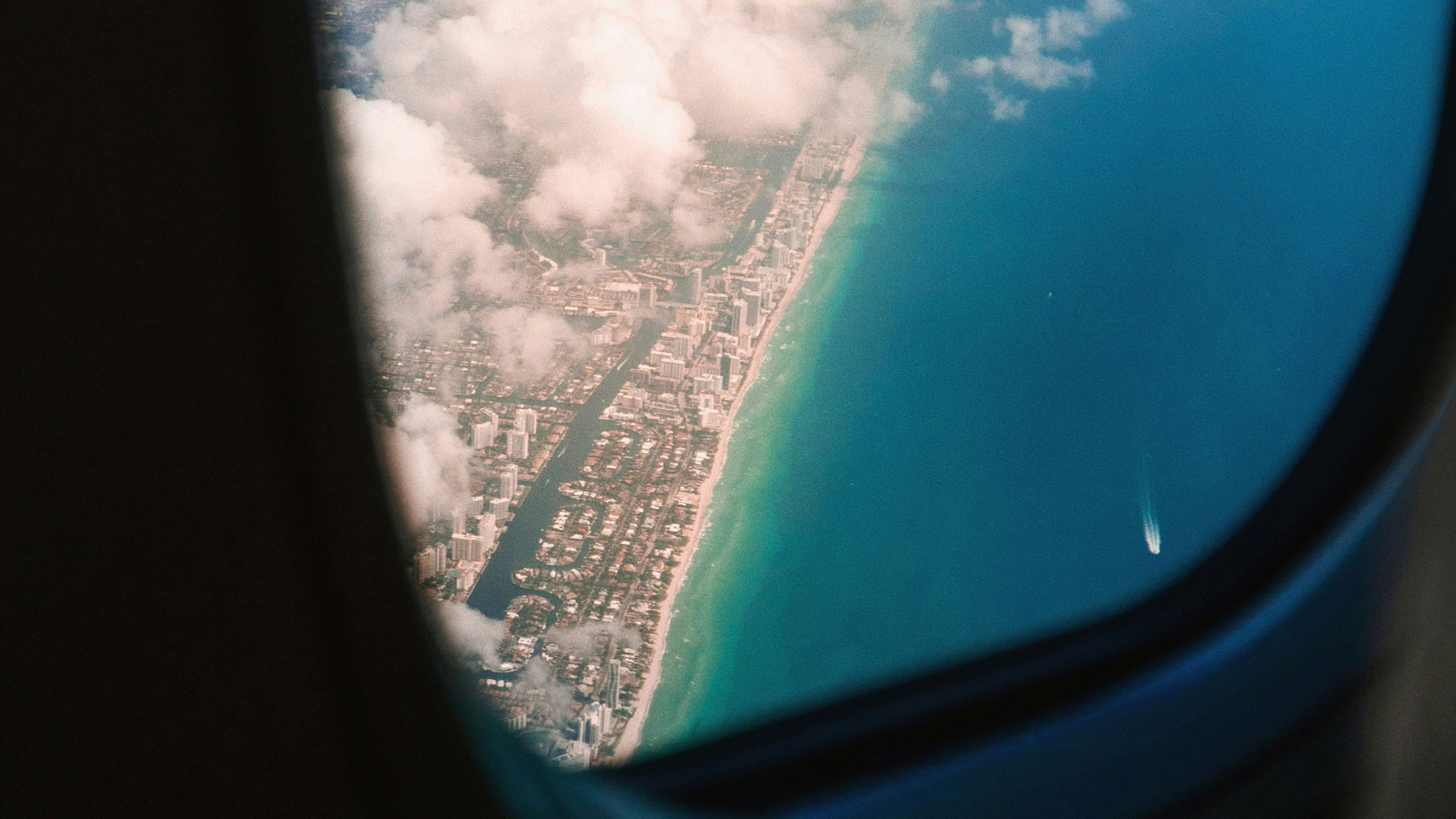 View from an airplane window showing a coastal city bordered by turquoise ocean waters and dotted with clouds.